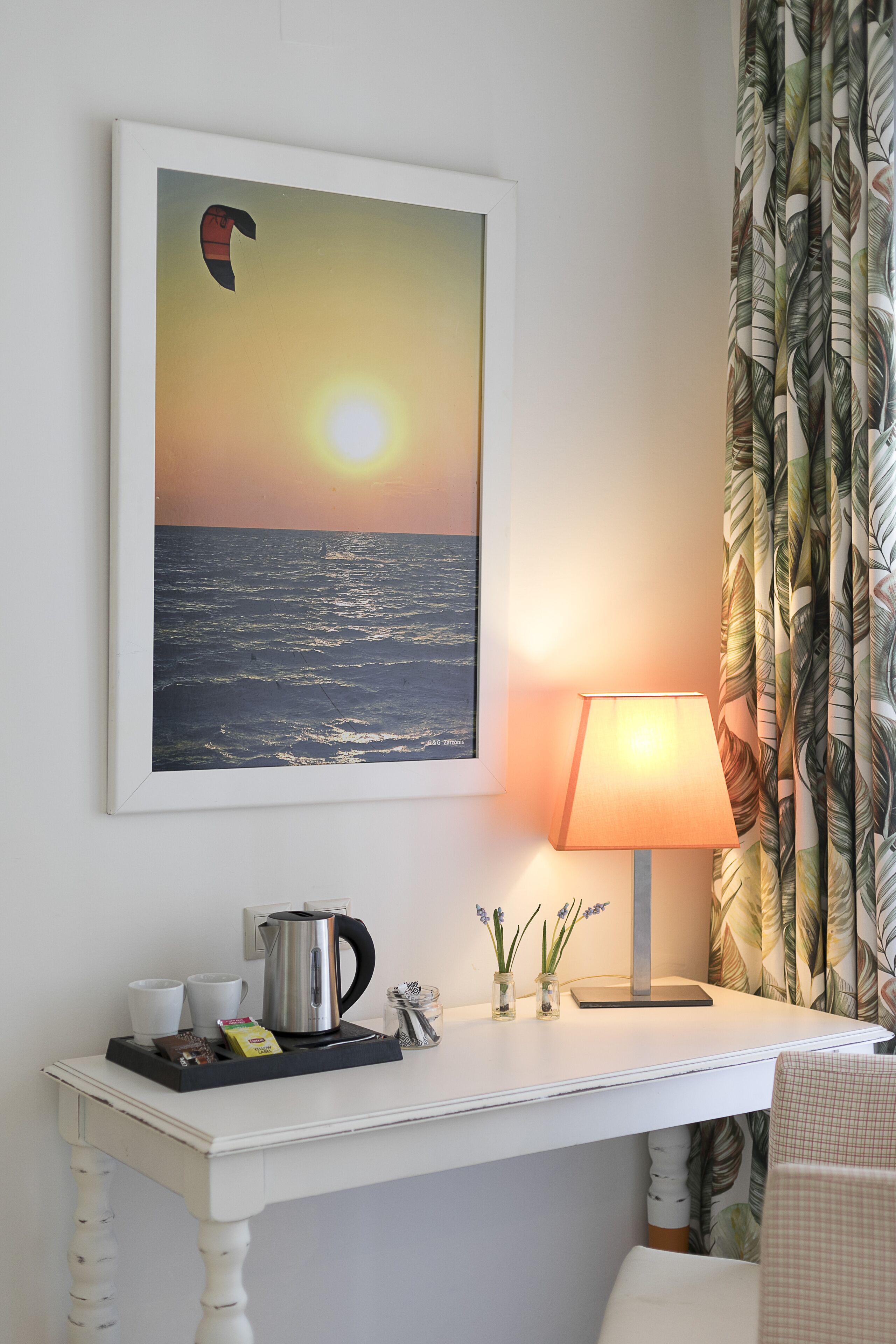 A framed sunset painting, a table with a lamp, a kettle, and a tray with cups and a box of tissues.