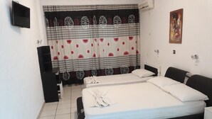 A double bed with white linens, a black headboard, and a bedside table. There is a TV mounted on the wall, a curtain with floral patterns, and a painting on the wall. - Skoufezis Studios (Kos)