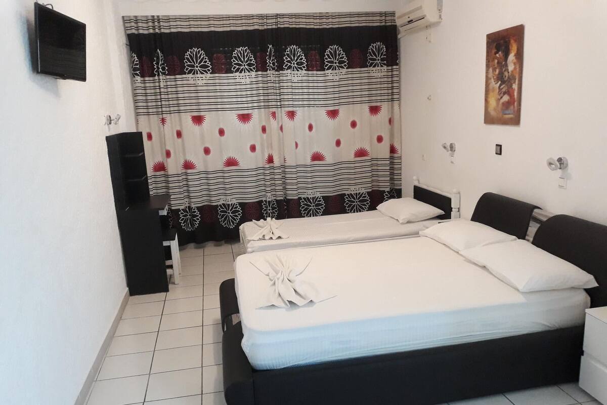 A double bed with white linens, a black headboard, and a bedside table. There is a TV mounted on the wall, a curtain with floral patterns, and a painting on the wall.