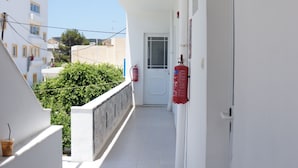 A narrow, white corridor with a fire extinguisher mounted on the wall, a door at the end, and a view of buildings and greenery outside. - Skoufezis Studios (Kos)