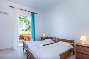 A clean, well-lit hotel room with two beds, a small table, and a chair near a window with a view of greenery. - Aparthotel Orestis (Corfu)