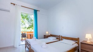 A room with two wooden beds, white linens, a small table with a chair, and a balcony with a view of greenery. - Aparthotel Orestis (Corfu)