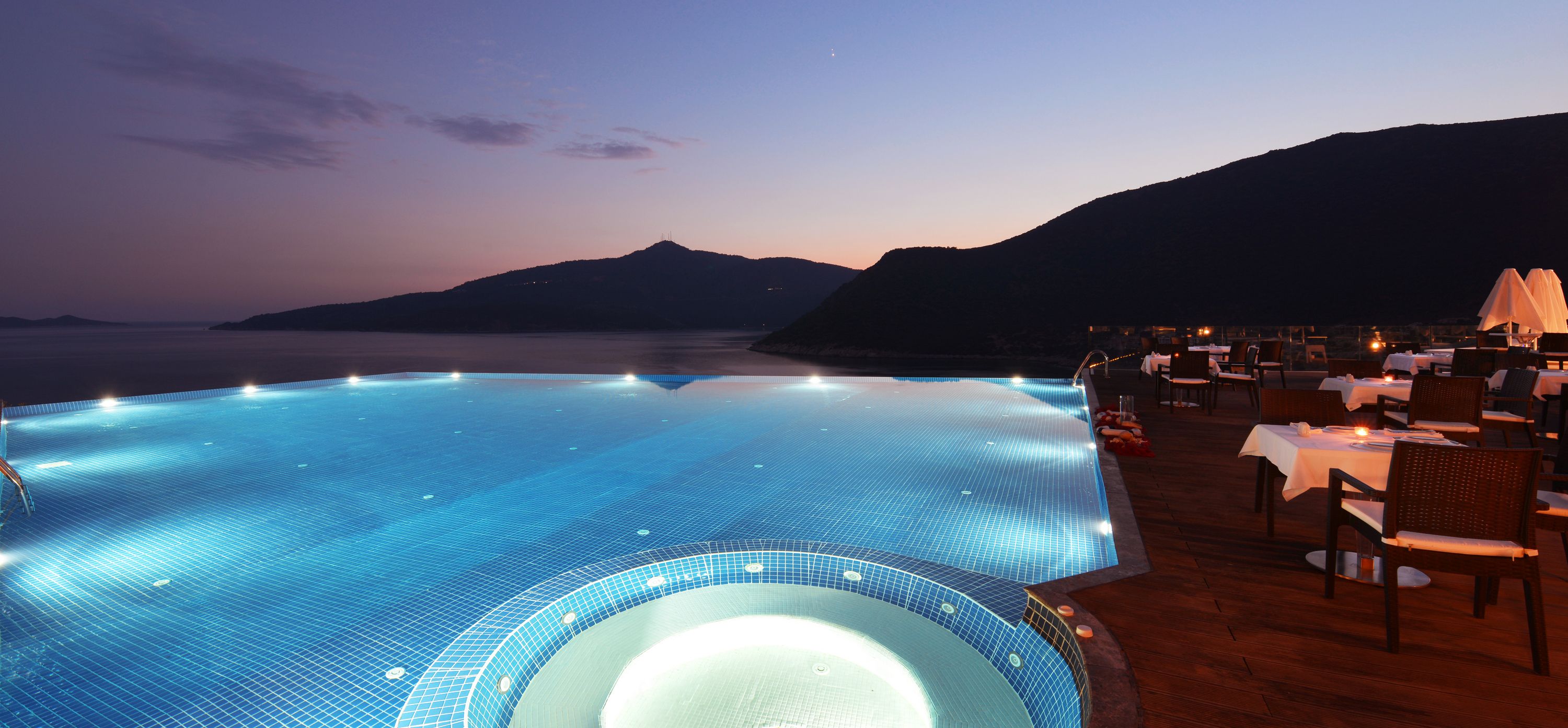 An infinity pool with a view of the sea and mountains at dusk.