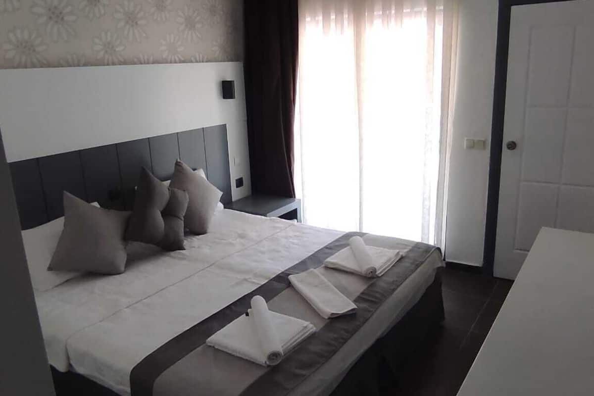 A modern hotel room with a large bed, a bedside table, a desk, and a wardrobe.