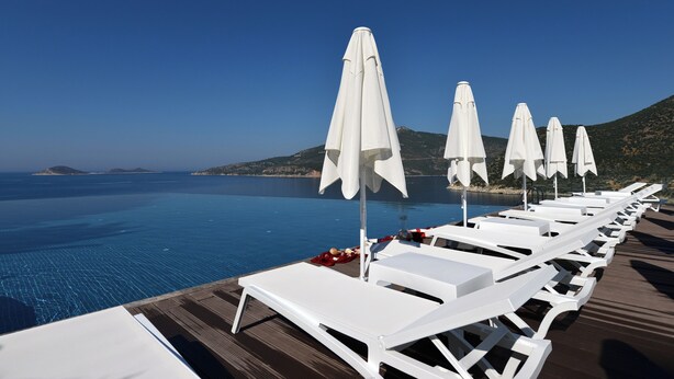 A row of white sun loungers with umbrellas, a wooden deck, and a clear view of the sea and hills in the background. - Kalamar Hotel (Kas)