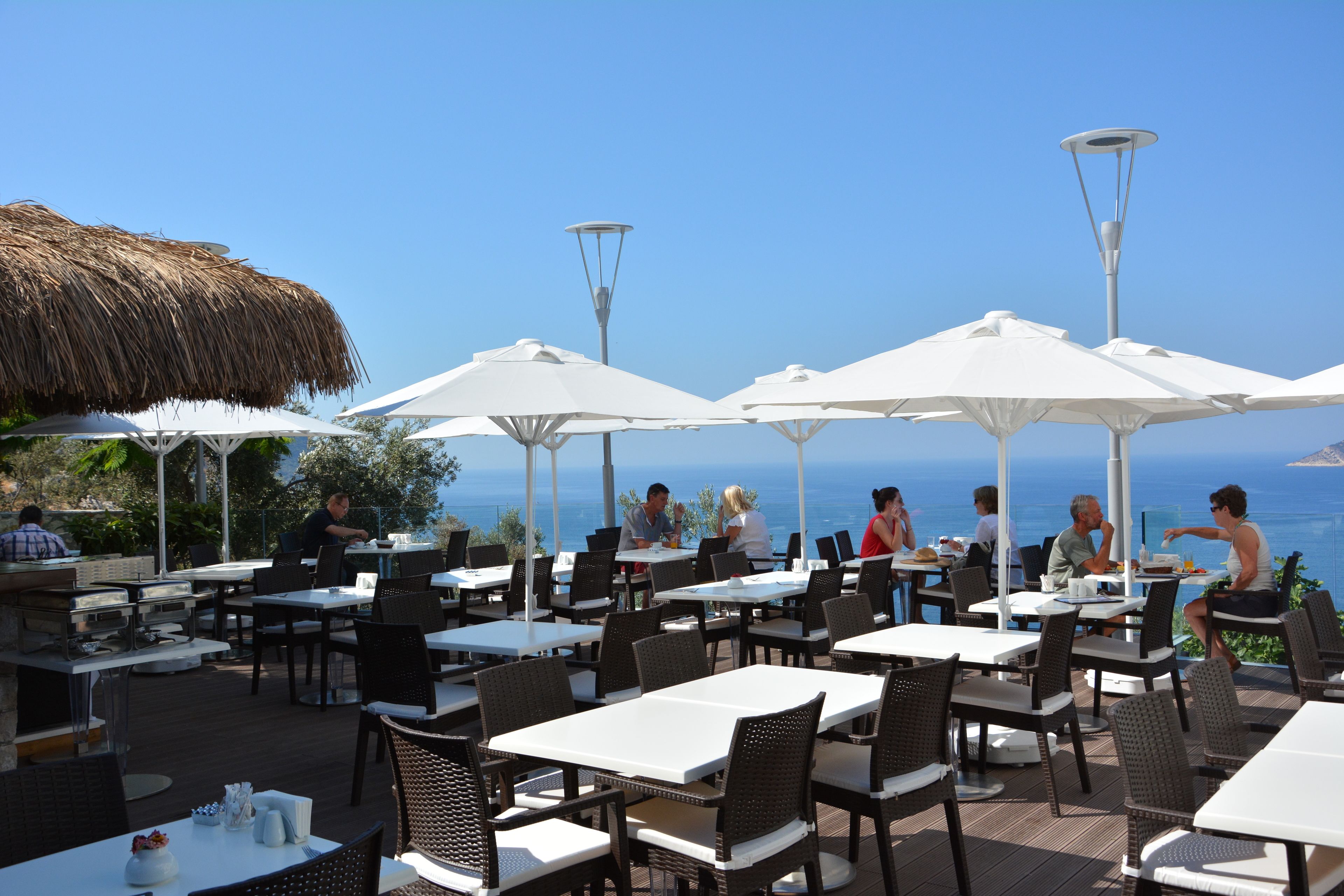 An outdoor dining area with tables and chairs, umbrellas, and a view of the sea.