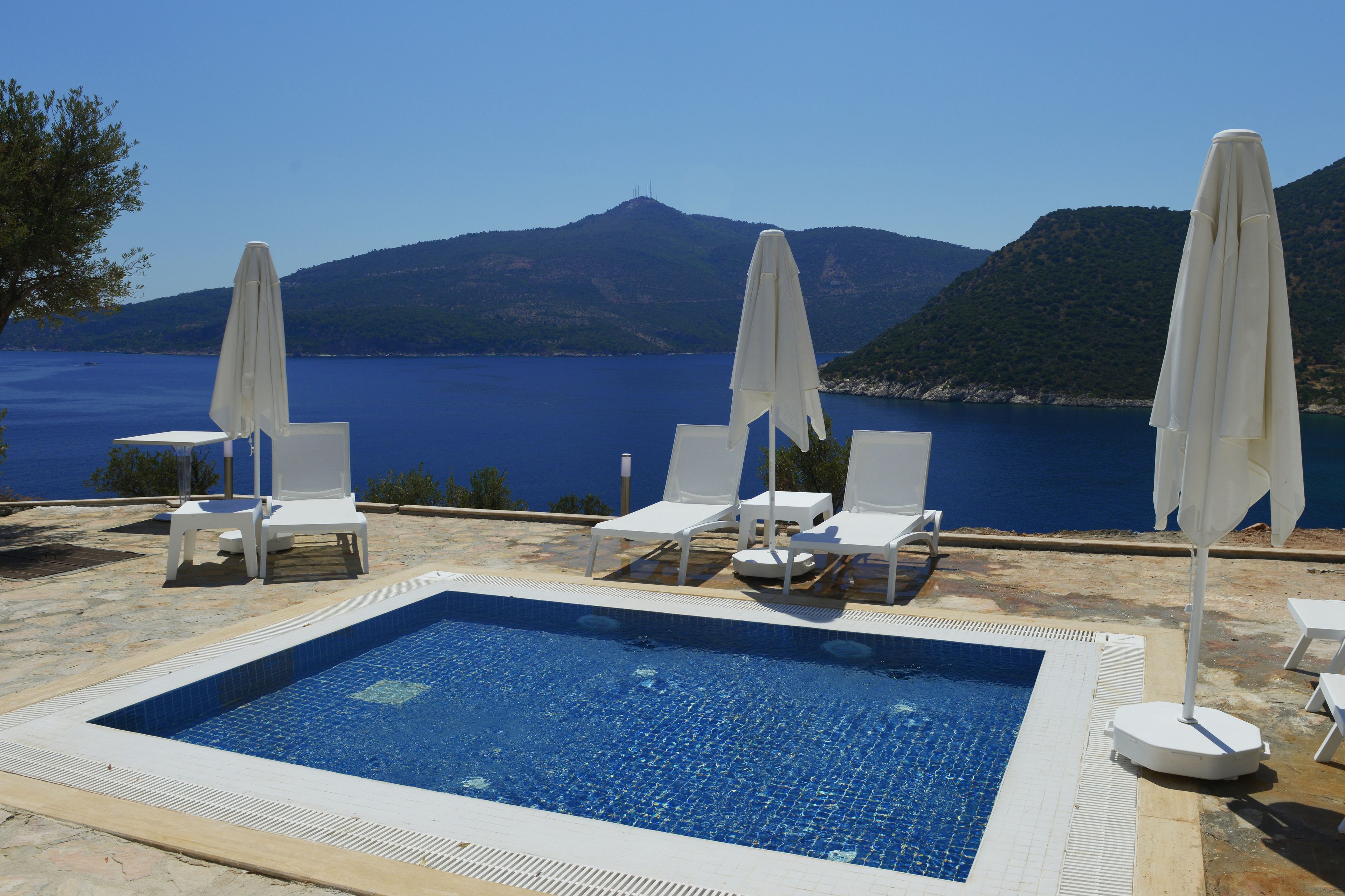 A pool with white lounge chairs and umbrellas, surrounded by a clear blue sea and hills.