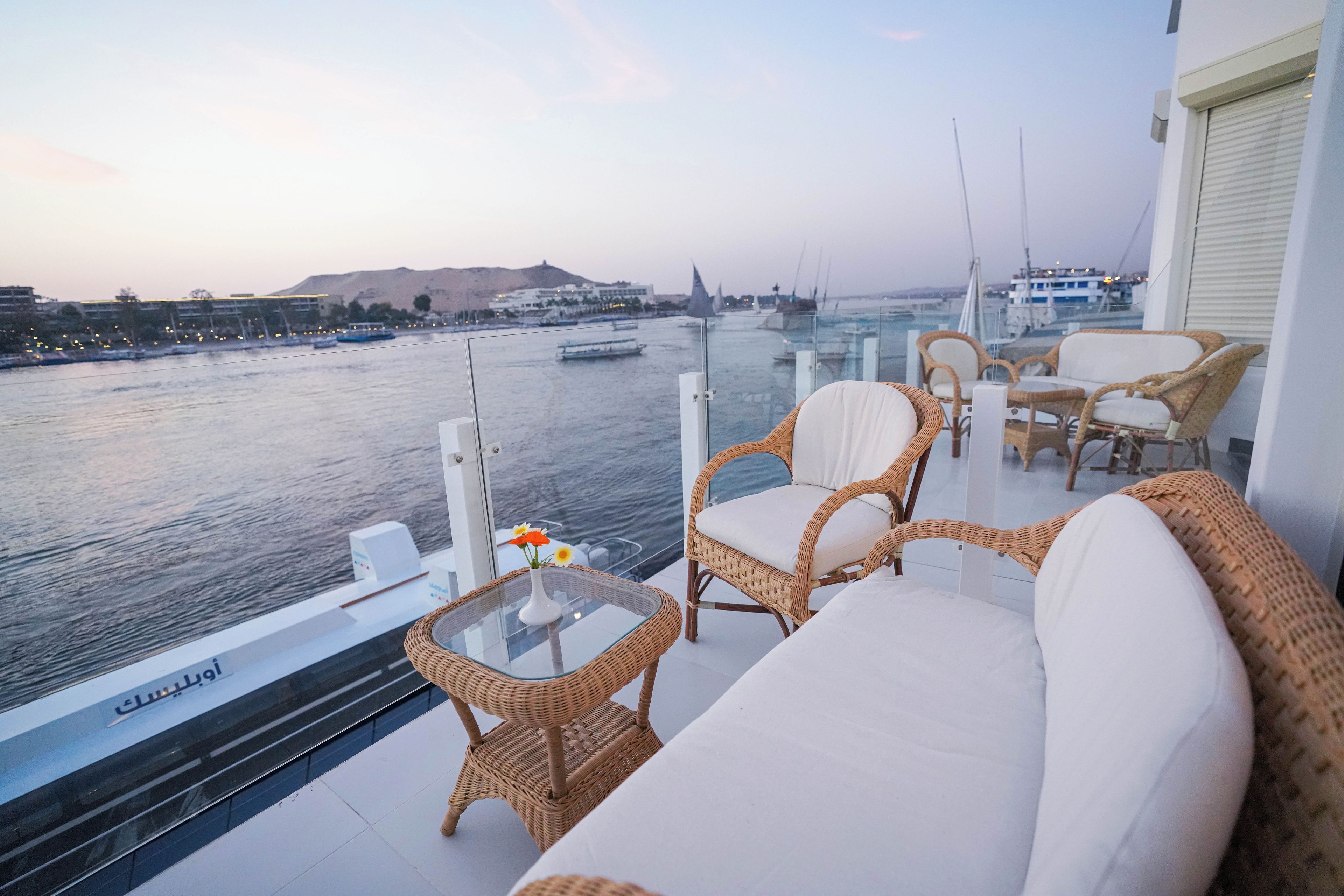 A balcony with wicker furniture, a glass table with a vase, and a view of the water with boats.