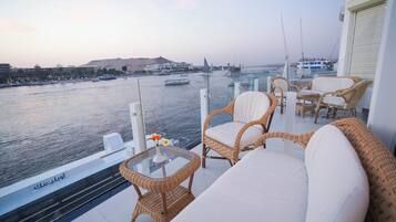 A balcony with wicker furniture, a glass table with a vase, and a view of the water with boats.