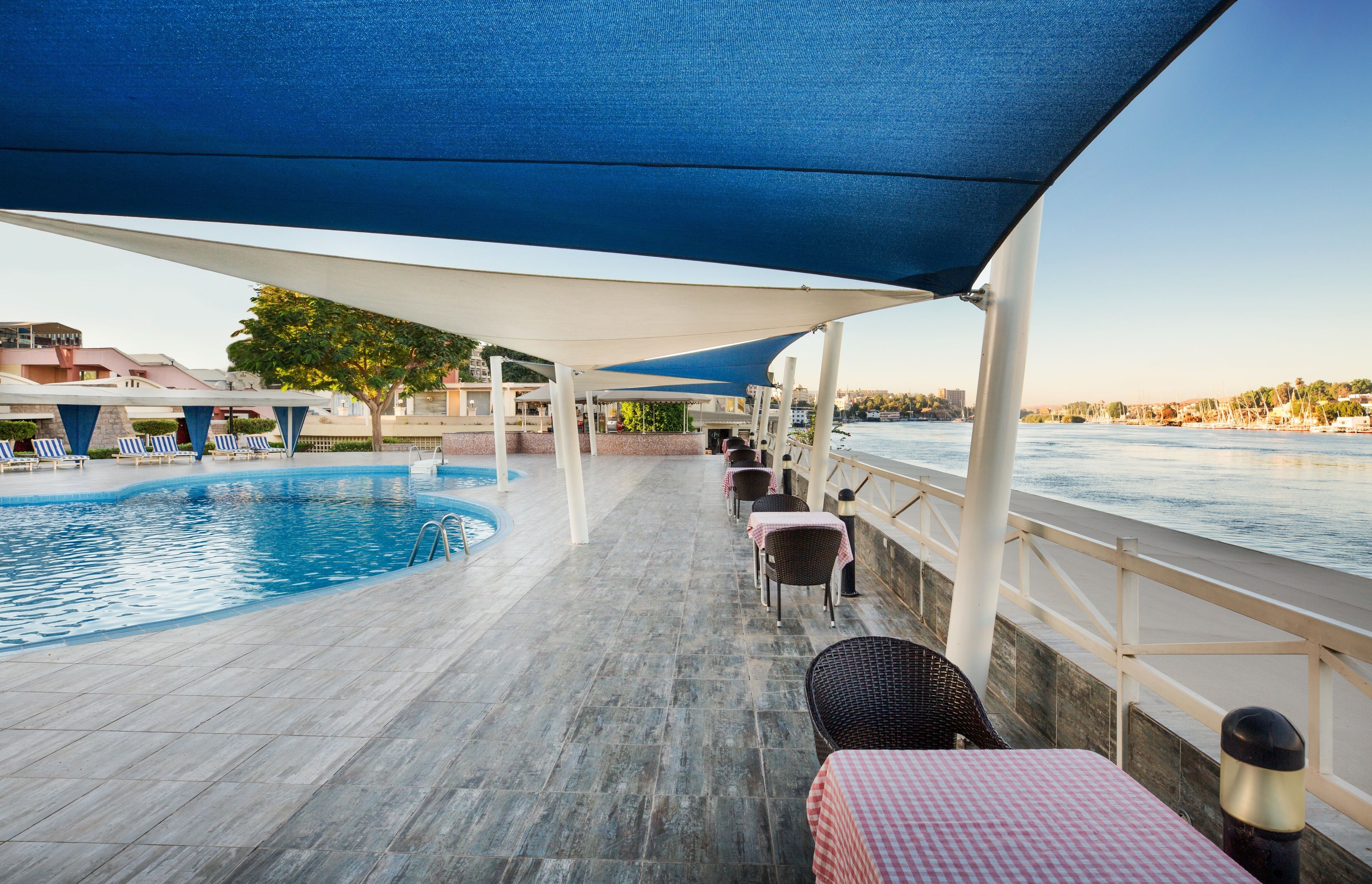Un espace aménagé au bord de la piscine, avec des chaises longues et des tables sous un auvent, donnant sur une étendue d’eau.