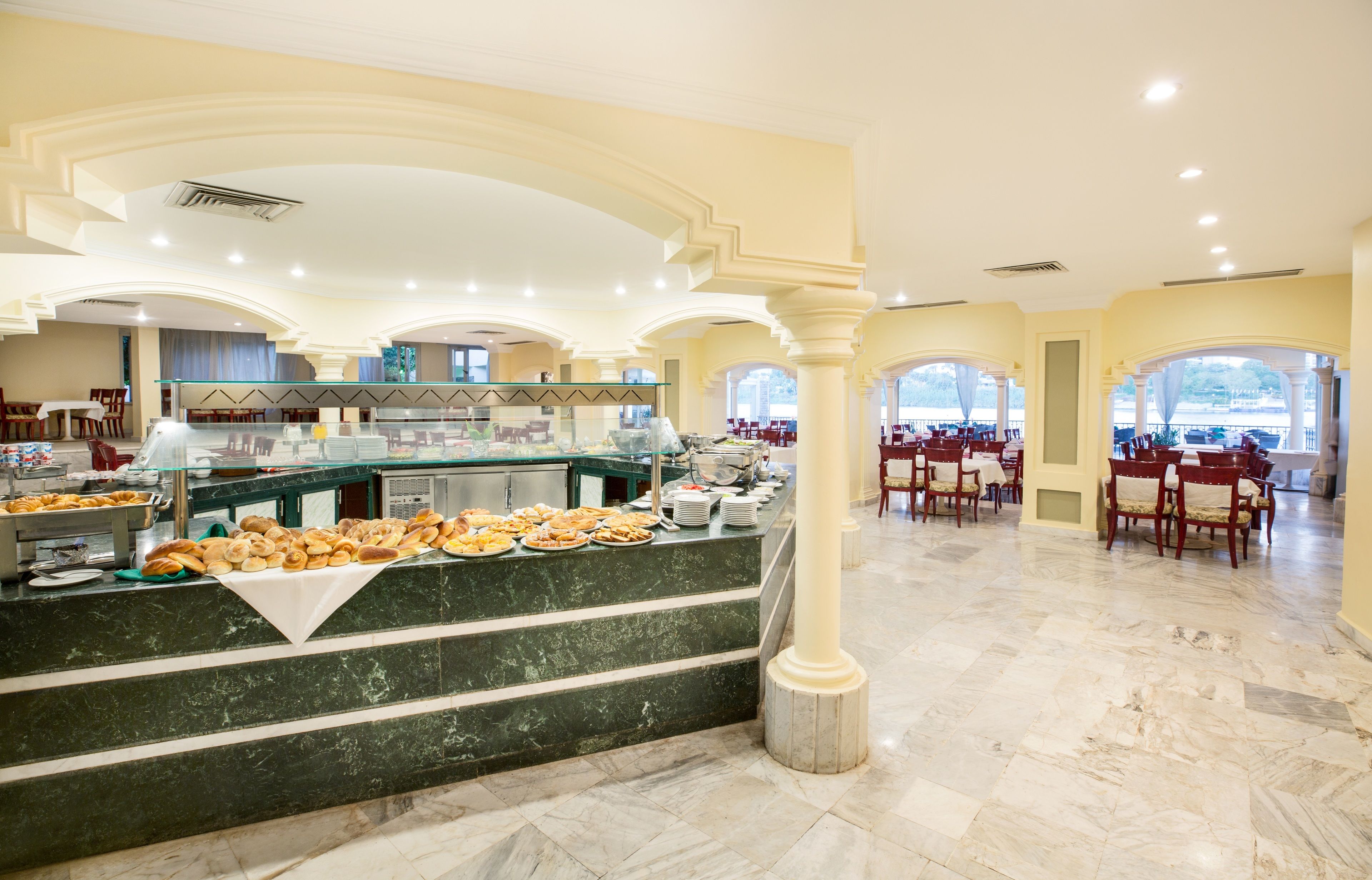 Zona de buffet con mostradores de mármol, diferentes opciones de comida y arreglos de asientos en un amplio salón con arcos y ventanas grandes.