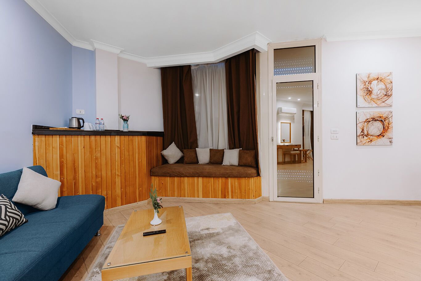 A modern living room with a blue sofa, wooden paneling, a small table with a vase, and a sliding glass door leading to a dining area.
