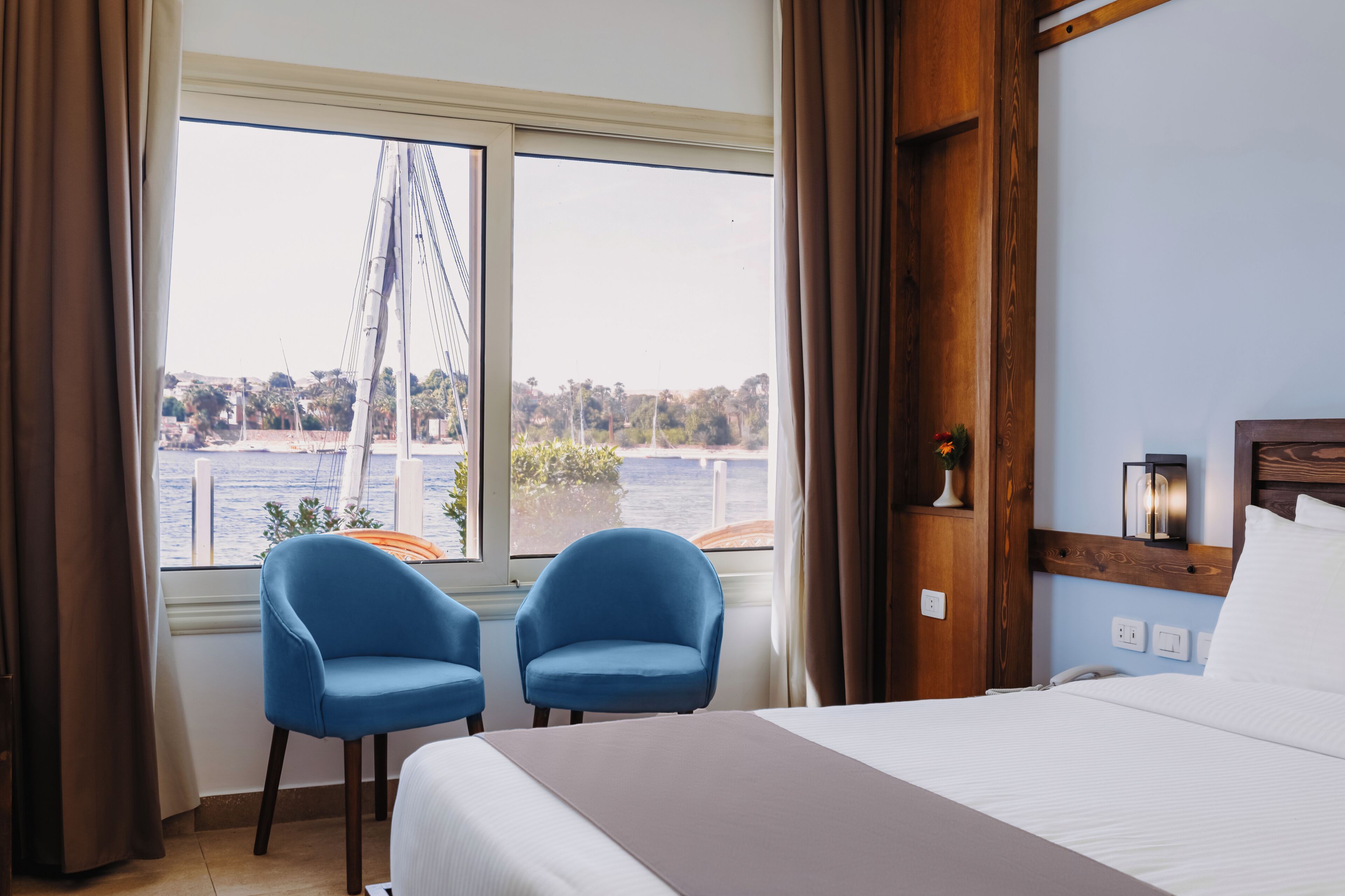 Ein Hotelzimmer mit einem Bett, zwei blauen Stühlen, einem Fenster mit Blick auf eine Brücke und Wasser, und einem hölzernen Kopfende.