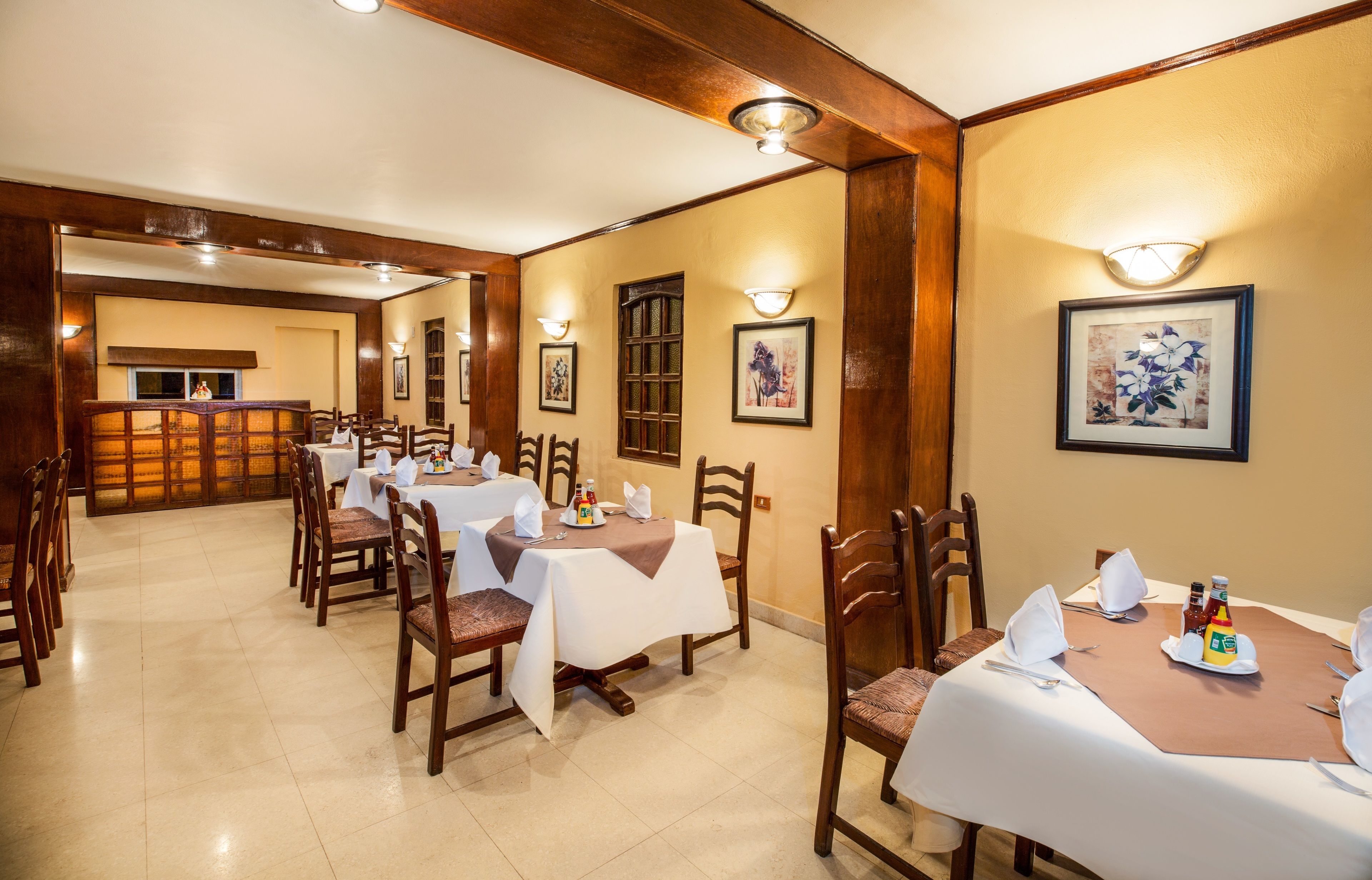 A dining area with tables set for four, wooden chairs, and wall-mounted light fixtures.