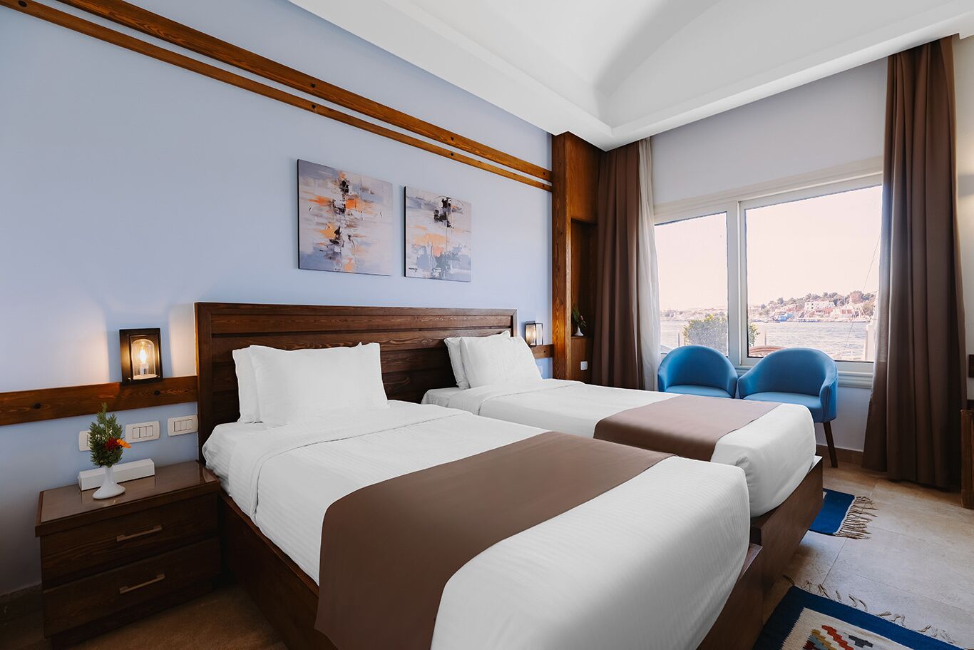 A hotel room with two beds, a nightstand, a lamp, a window with a view, and two blue chairs.
