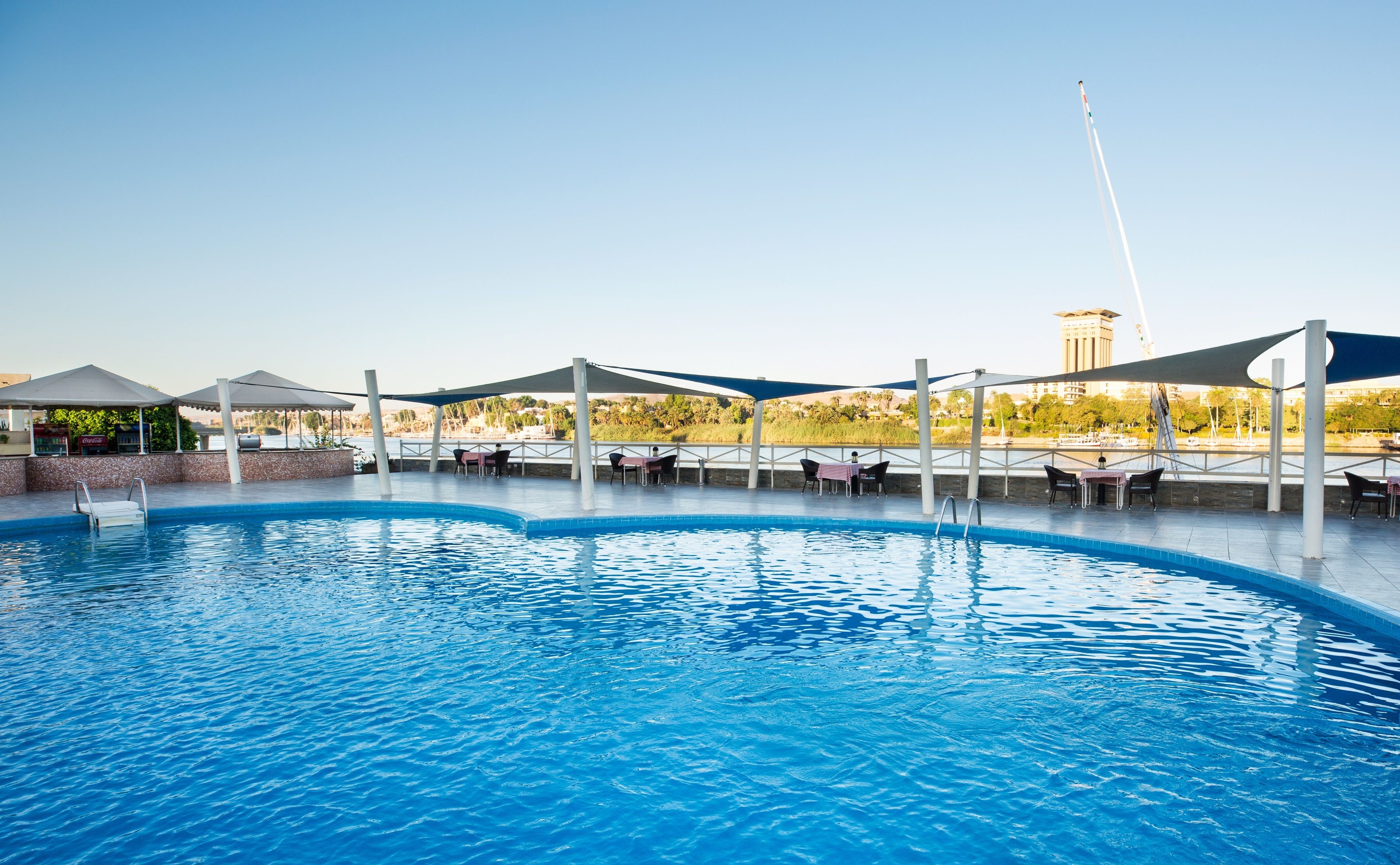 A swimming pool with lounge chairs and umbrellas, surrounded by a shaded area with tables and chairs.