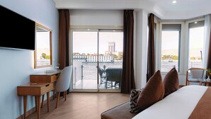 Habitación de hotel con cama, escritorio, silla y un ventanal con vistas a un edificio y una masa de agua.