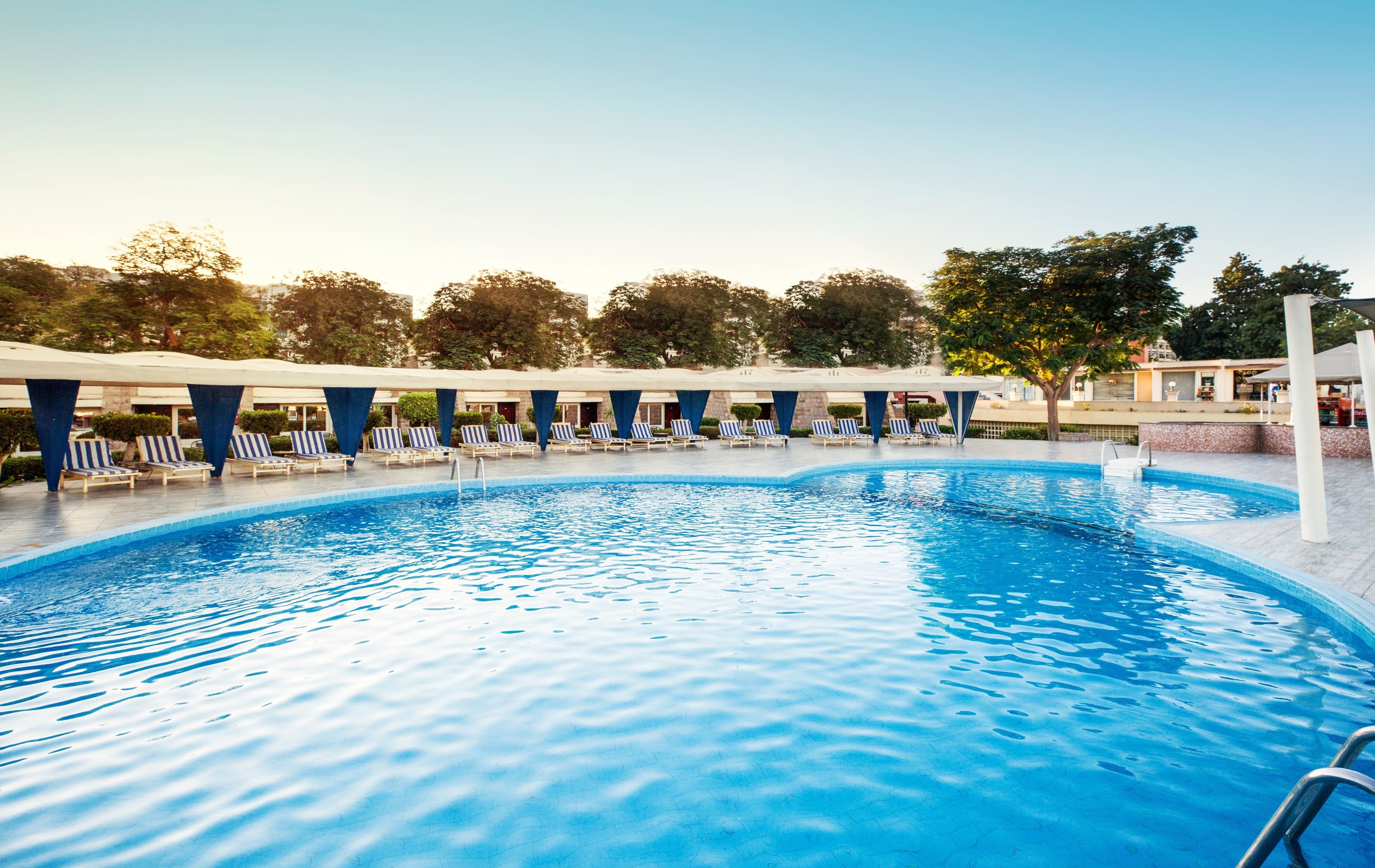 A swimming pool with lounge chairs and umbrellas, surrounded by trees and buildings.