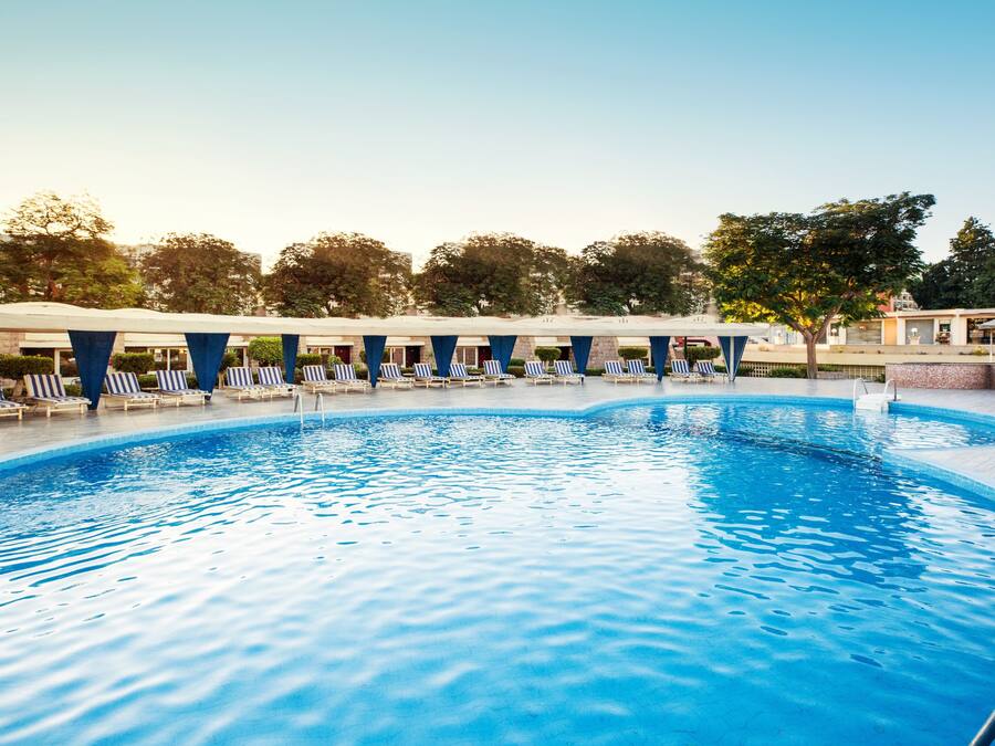 A swimming pool with lounge chairs and umbrellas, surrounded by trees and buildings.