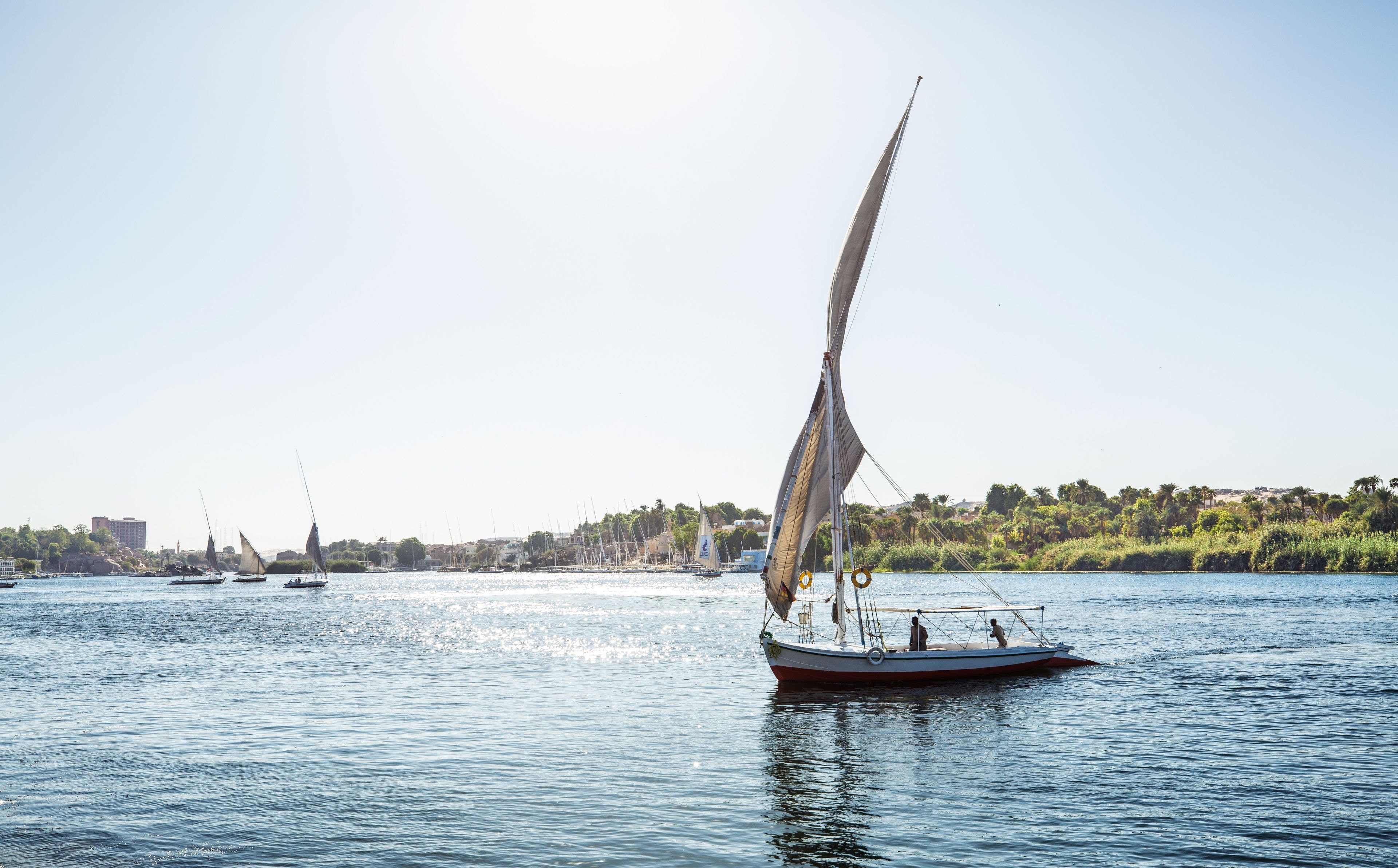 A sailboat on calm waters with a distant shoreline of buildings and greenery.