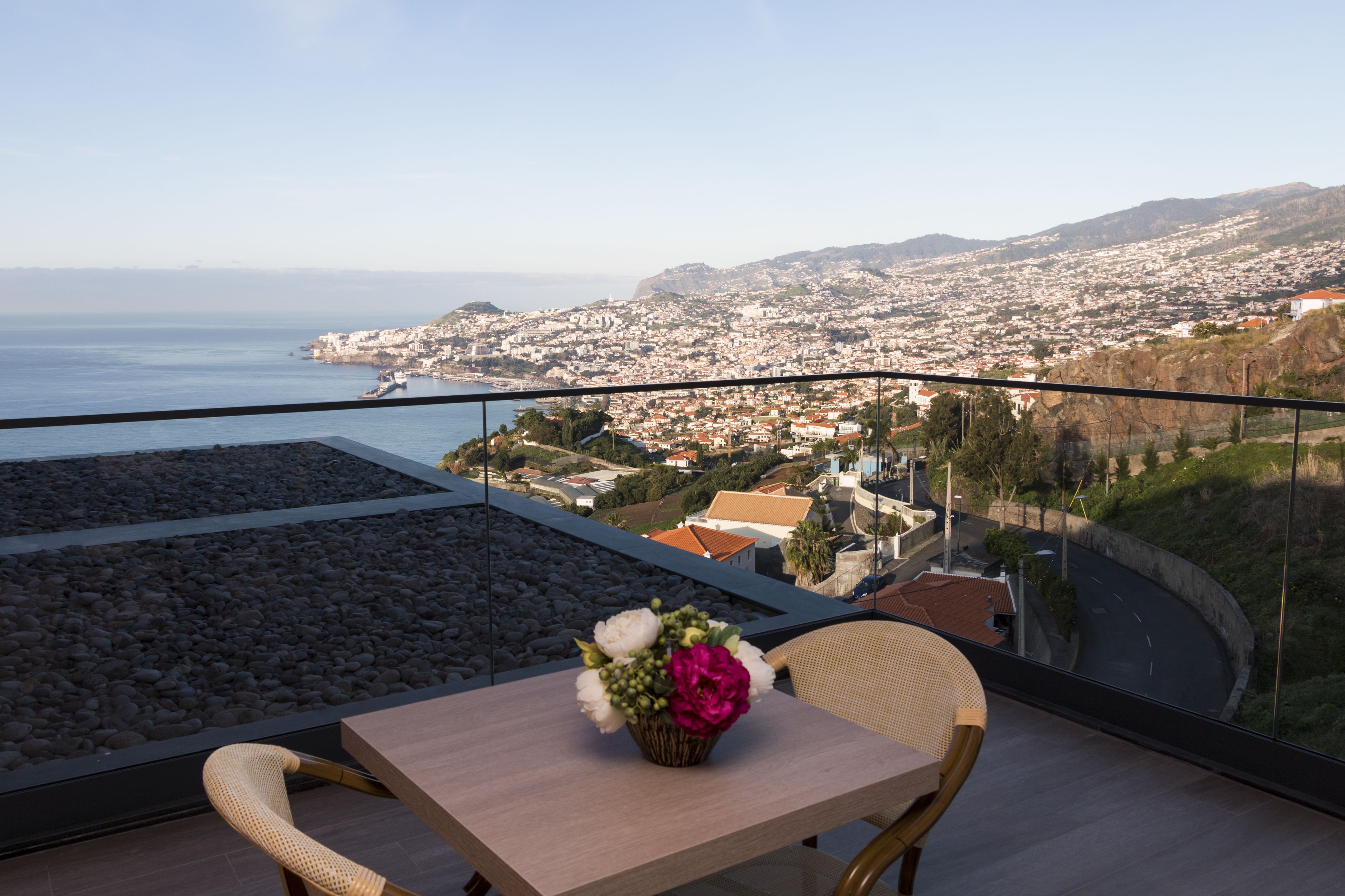Ein Balkon mit Tisch und Stühlen, einer Blumenvase und Blick auf eine Küstenstadt und Berge.
