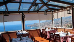 A restaurant with tables set for dining, large windows offering a view of the sea and city, and hanging lanterns. - Hotel Ocean Gardens (Funchal)