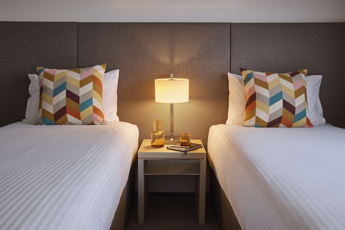 Habitación de hotel con dos camas, cada una con un cojín estampado, una mesita de noche con lámpara y una botella.