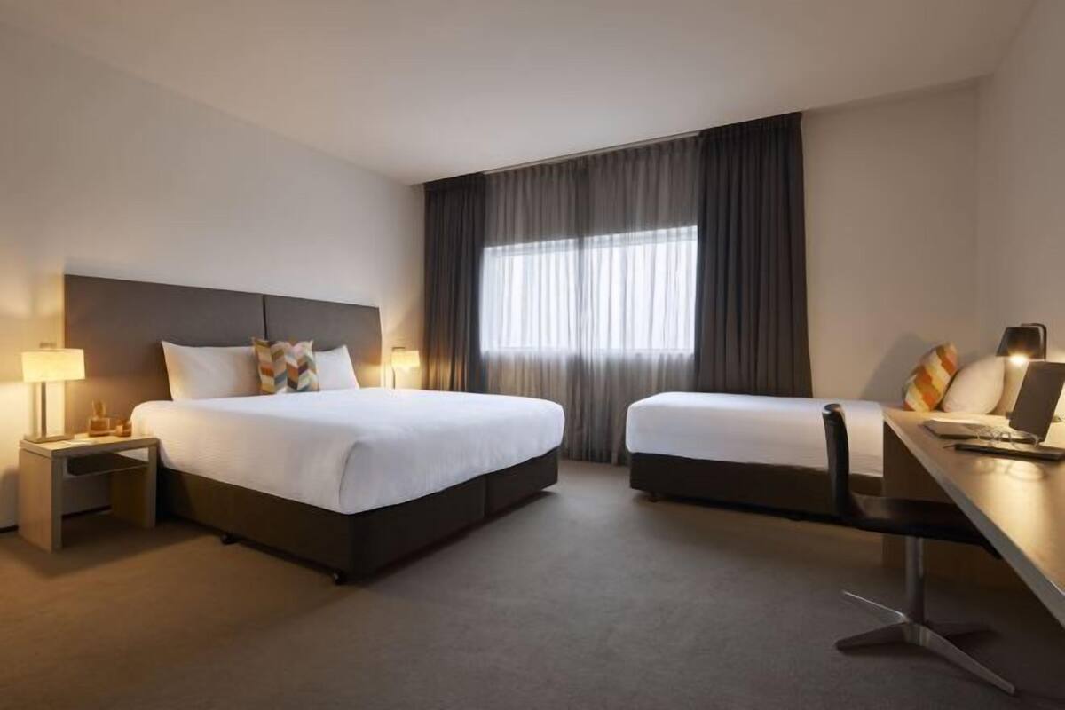 Habitación de hotel con una cama grande, una cama más pequeña, un escritorio con silla, una mesita de noche y una ventana con cortinas.