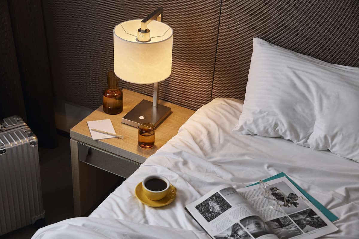 Una cama bien hecha con sábanas blancas, una lámpara de noche, una taza de café y una revista sobre la cama.
