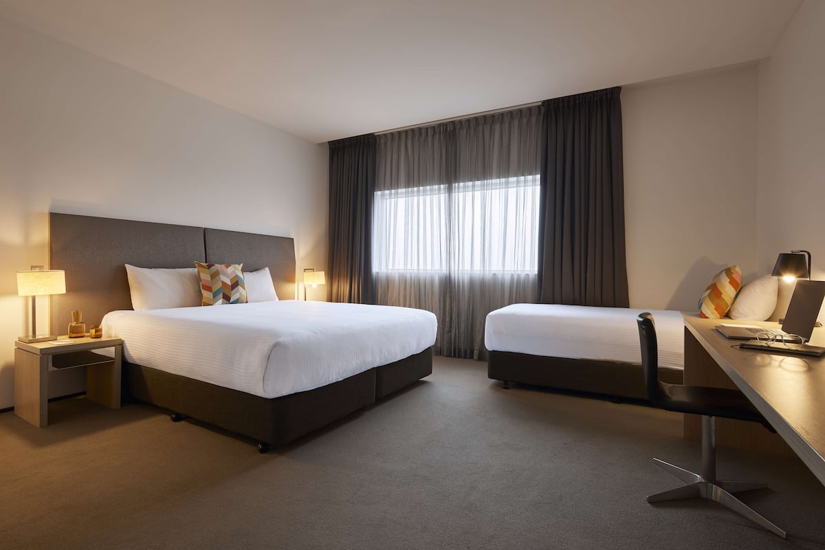 Habitación de hotel con una cama grande, una cama más pequeña, un escritorio con una silla, una lámpara y una ventana con cortinas.