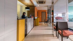Een hotel receptie met een baliemedewerker, een receptiebalie, een zithoek met stoelen en uitzicht op de straat.