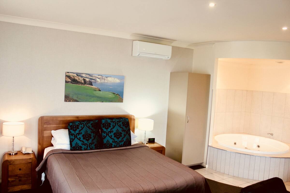Una habitación de hotel con una cama, lámparas de noche, un cuadro, un baño con bañera y un equipo de aire acondicionado.