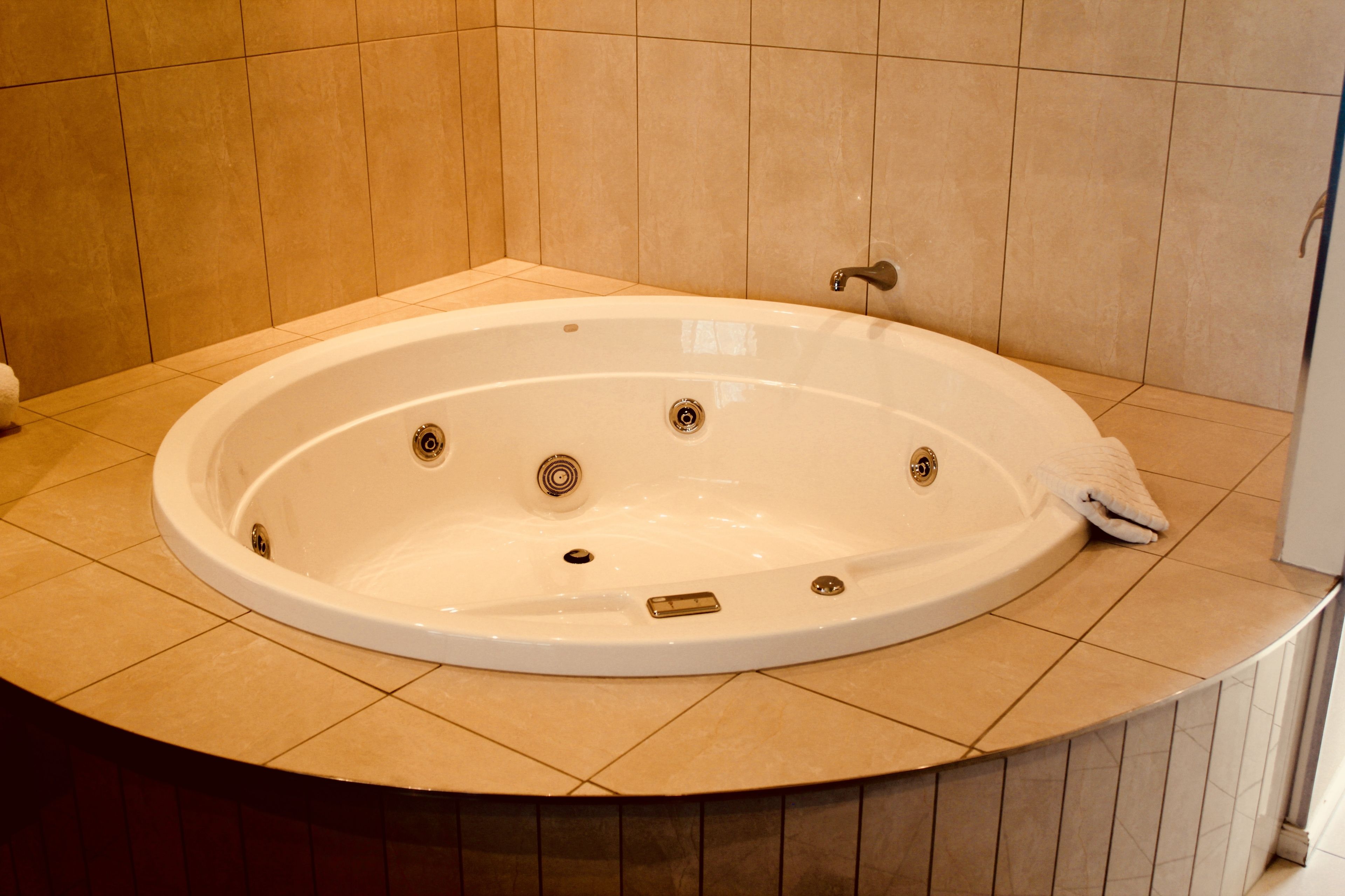 Uma banheira grande e oval, com uma única torneira e ralo, instalada em um canto com paredes e piso revestidos de azulejos bege.