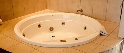 A large, oval-shaped hot tub with a single faucet and a drain, set in a corner with beige tiled walls and floor.