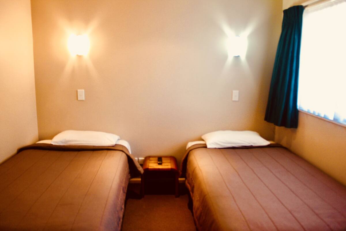 Un alojamiento pequeño y de una sola habitación con dos camas individuales, una mesita de noche y una lámpara fijada a la pared.
