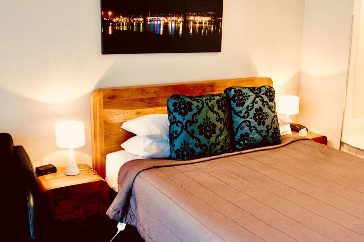 Habitación de hotel con cabecera de madera, dos lámparas de noche, cama con manta marrón y vista nocturna de un paisaje urbano.