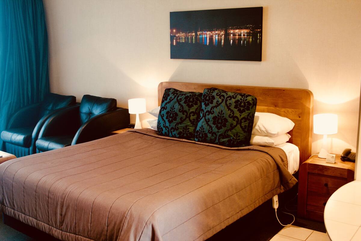 Habitación de hotel con una cama grande, dos lámparas de noche, una mesita de noche, una silla y un cuadro de un paisaje urbano en la pared.
