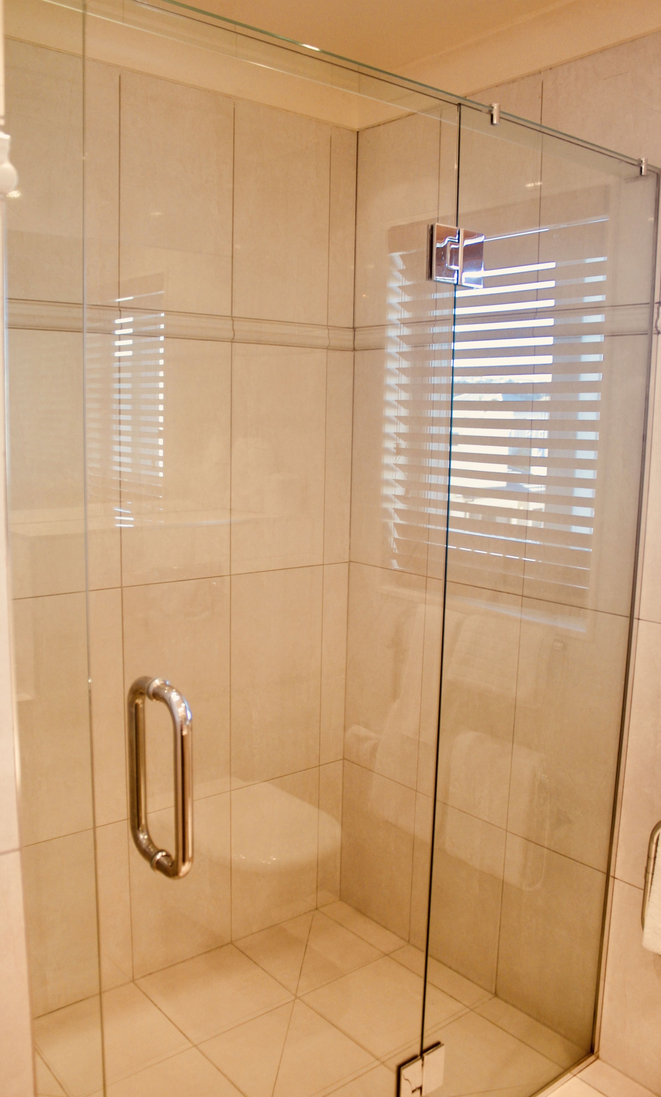 A glass-enclosed shower stall with a metal handle, tiled walls, and a window with blinds.