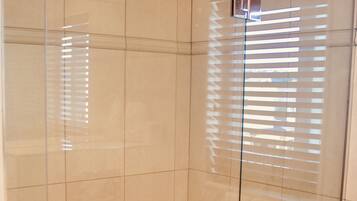 A glass-enclosed shower stall with a metal handle, tiled walls, and a window with blinds.