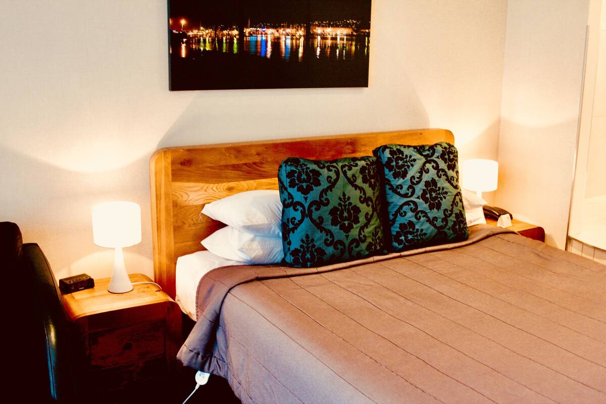 Una cama bien tendida con cabecera de madera, dos lámparas de noche y una mesita con lámpara.