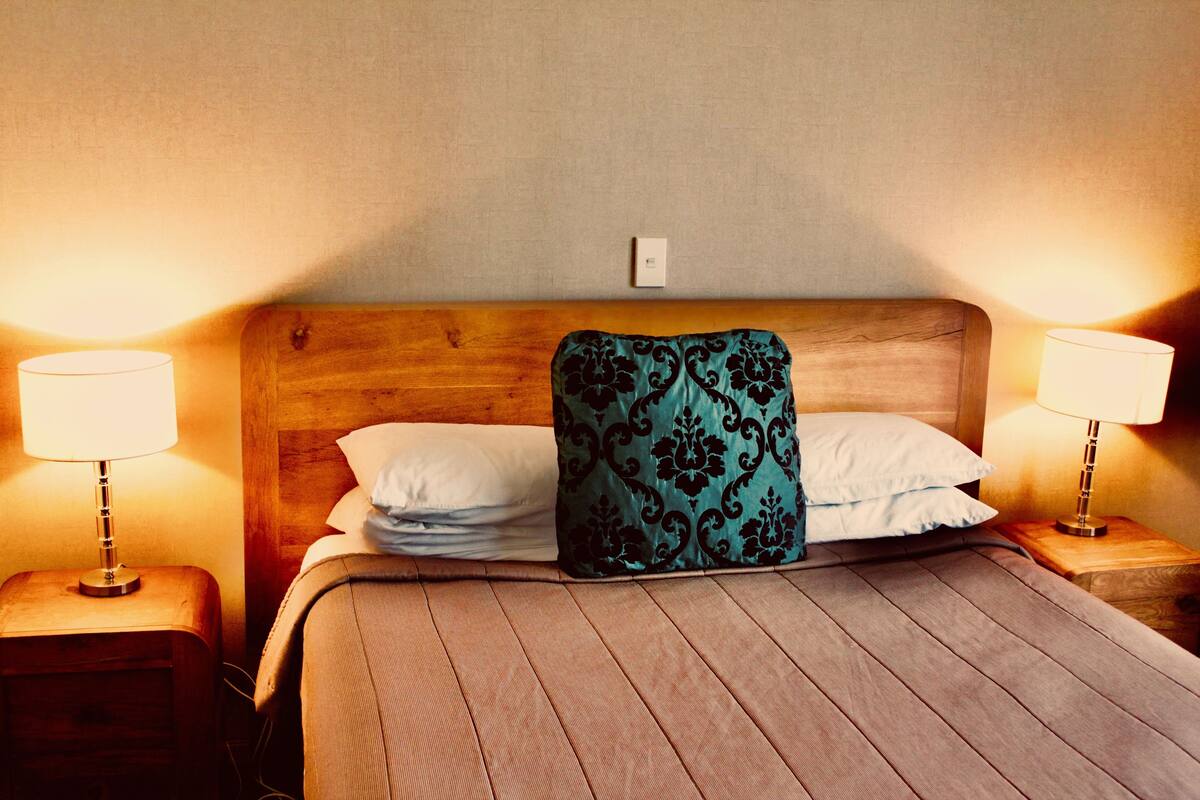 Una cama bien hecha con un cabecero de madera, dos lámparas de noche y un enchufe en la pared.