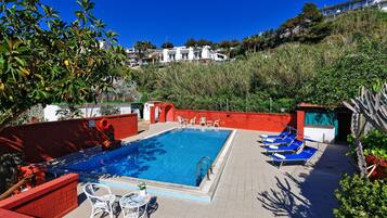 Piscina rectangular con solárium de baldosas, rodeada de muros rojos y algunas sillas de descanso.
