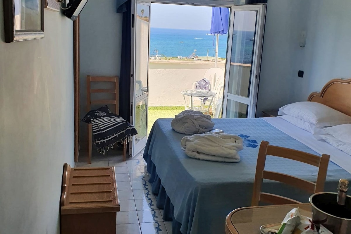 A room with a bed, a chair, a table with snacks, and a view of the beach through an open door.