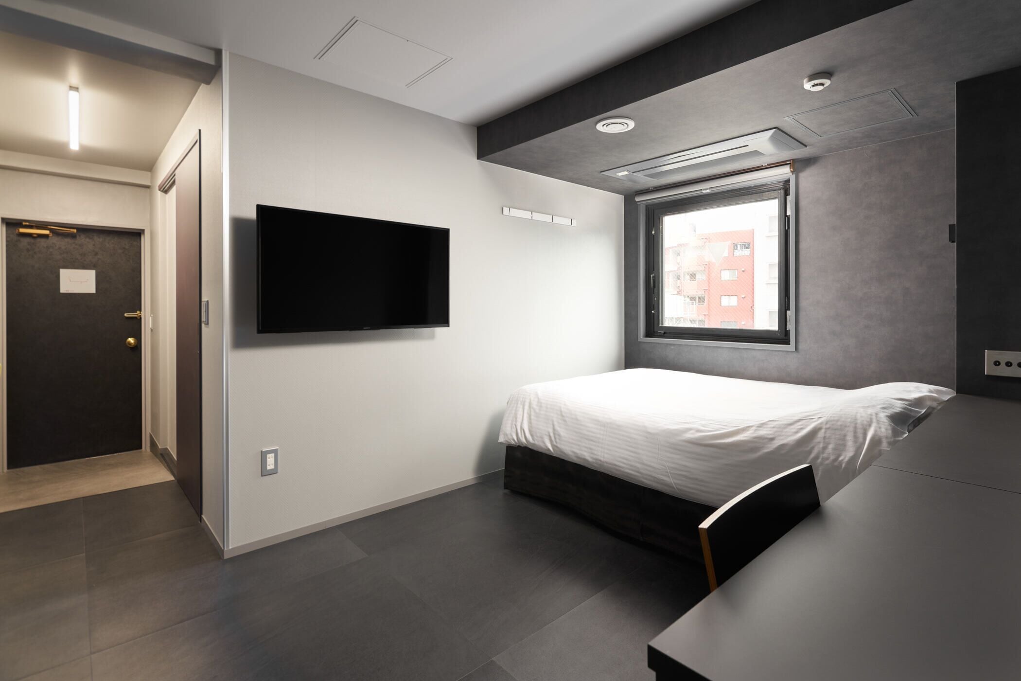 A modern hotel room with a bed, a flat-screen TV mounted on the wall, a window with a view of buildings, and a door with a handle.