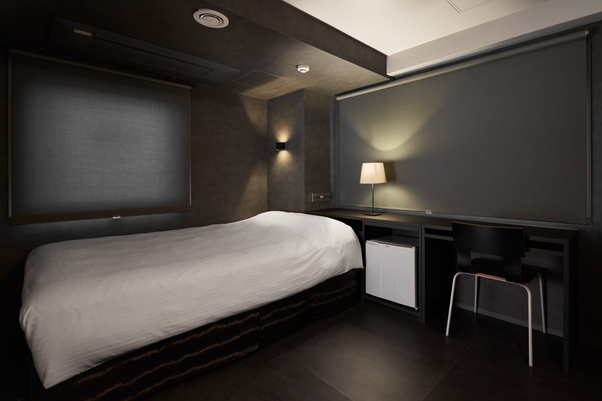 A modern hotel room with a bed, desk, chair, and a wall-mounted lamp.