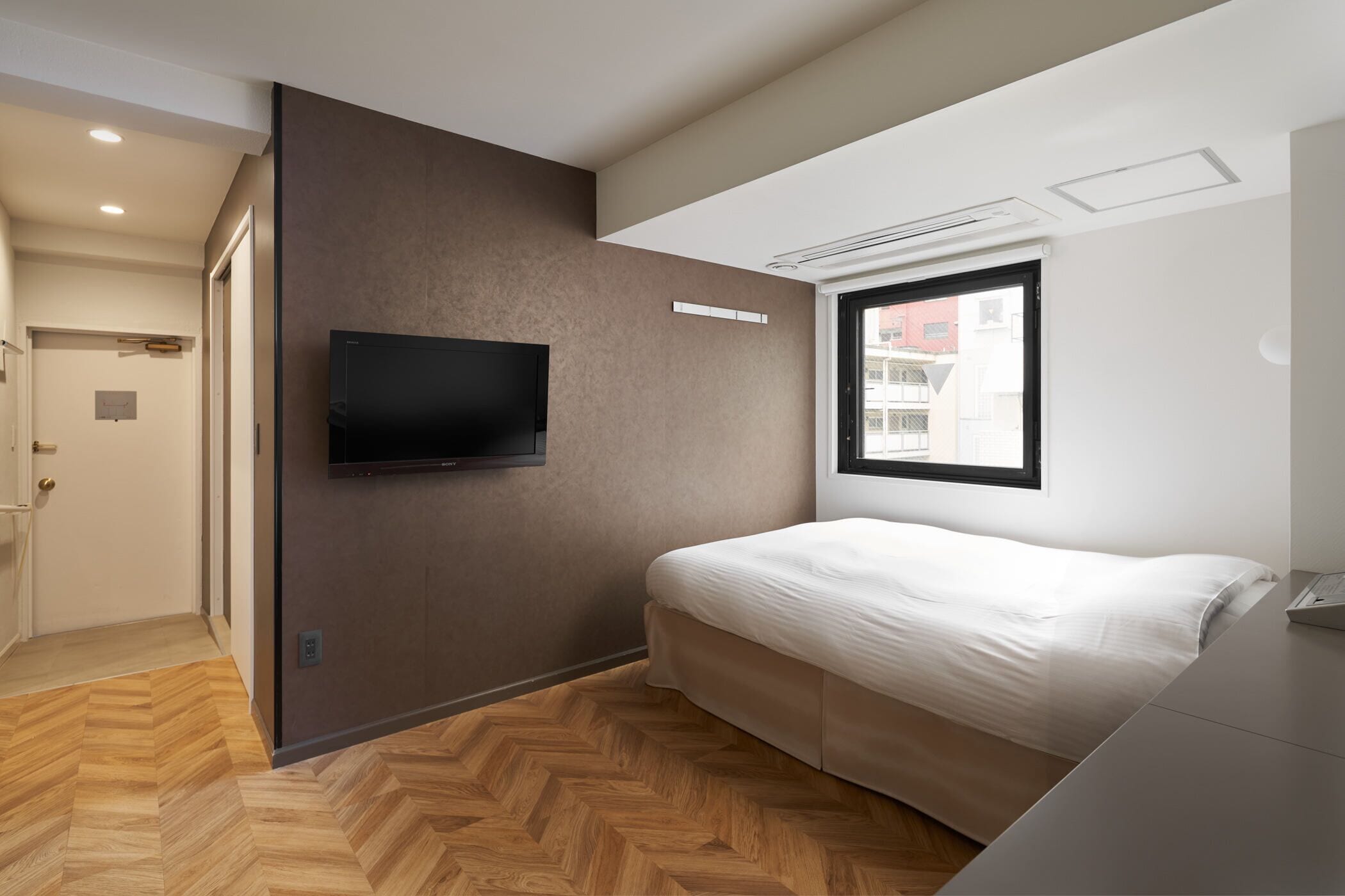 A modern bedroom with a bed, a flat-screen TV mounted on a wall, a window with a view of buildings, and a wooden herringbone floor.