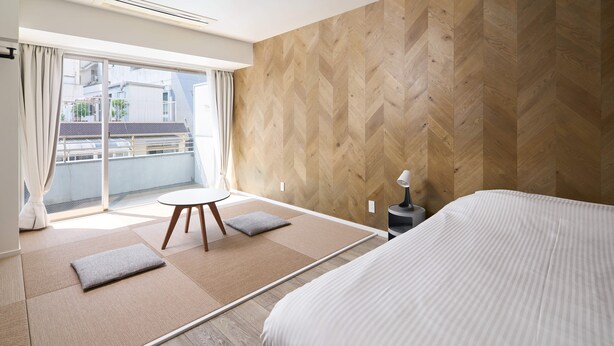 A modern hotel room with a large bed, a small round table, a sofa, and a view of the city through large windows. - City Hotel N.U.T.S. (Tokyo)