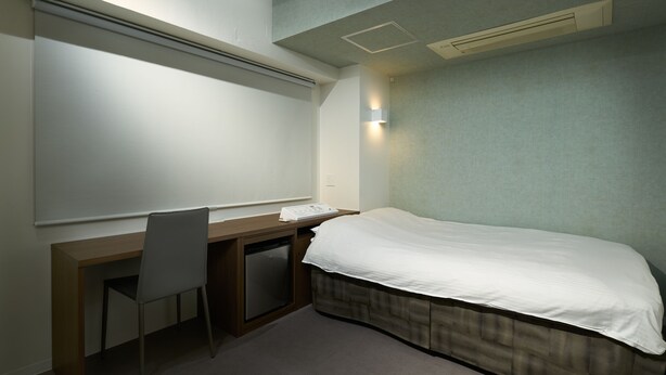 A hotel room with a bed, a desk, a chair, a small bedside table, and a wall-mounted air conditioning unit. - City Hotel N.U.T.S. (Tokyo)