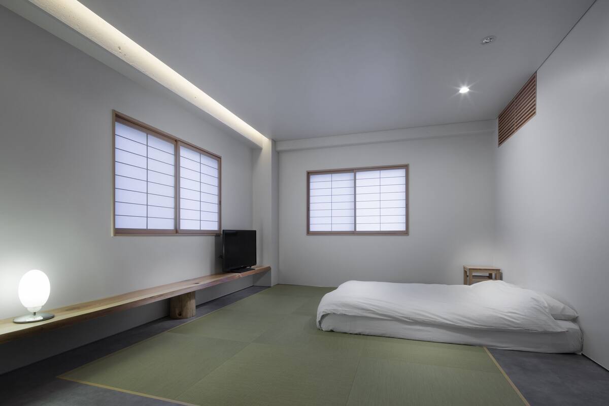 A minimalist room with tatami flooring, a bed, a TV on a low shelf, and two windows with wooden frames.