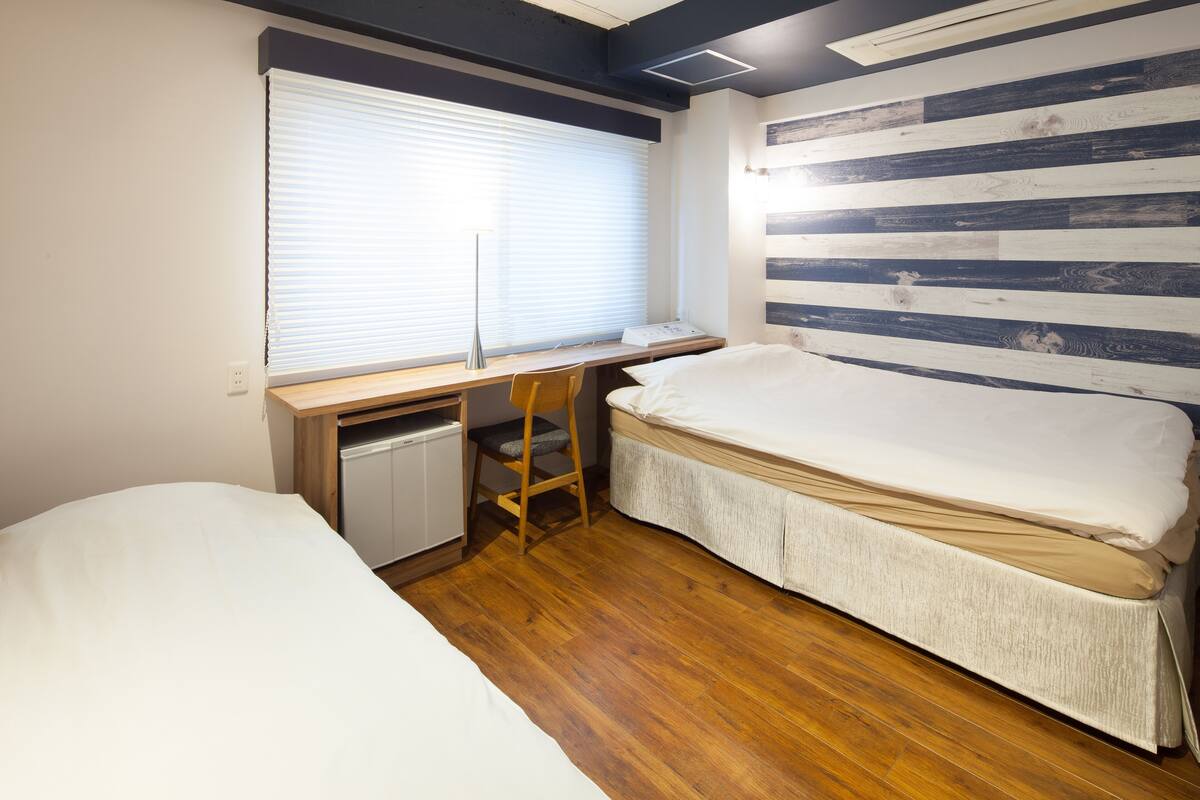 A bedroom with a wooden floor, a bed, a desk with a chair, and a window with blinds.